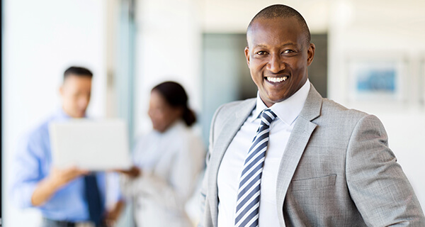 A business man in a gray suit and blue-striped tie smiles and stands tall.