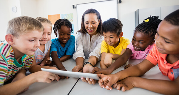 A teacher and five young students stand around a table looking at something displayed on an iPad. 