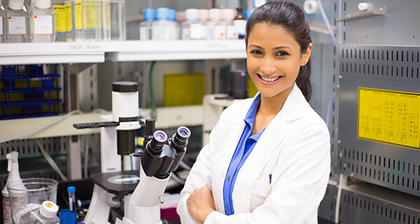 A female health science professional in a white lab coat stands next to a microscope.