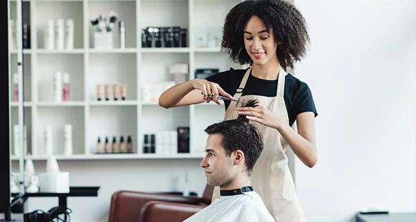 A female hair stylist gives a young man in a chair a hair cut.